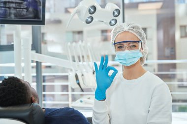 Gözlüklü ve maskeli bir doktor hastanın yanında oturuyor ve stomatolojide çalışmaya hazırlanıyor, kameraya bakıyor.