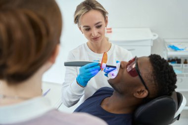 Koruyucu eldivenli kadın doktor erkeğin yanında duruyor ve dişçide ultrasonik diş temizliği yapıyor.