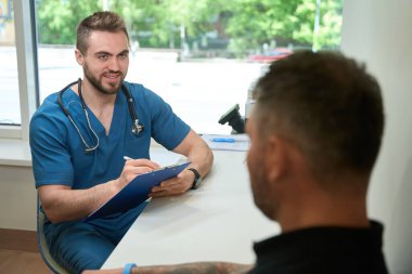 Gülümseyen tıp doktoru masasına oturmuş hasta şikayetleri yazıyor.