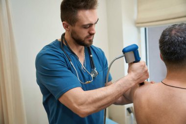 Erkek hastanın omzunda ekstrakorporeal şok terapi cihazını yöneten odaklı fizik terapist.