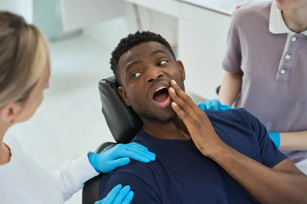 椅子に座って看護師を見ている歯痛の若い患者 男性に話す保護手袋の医師 — ストック写真