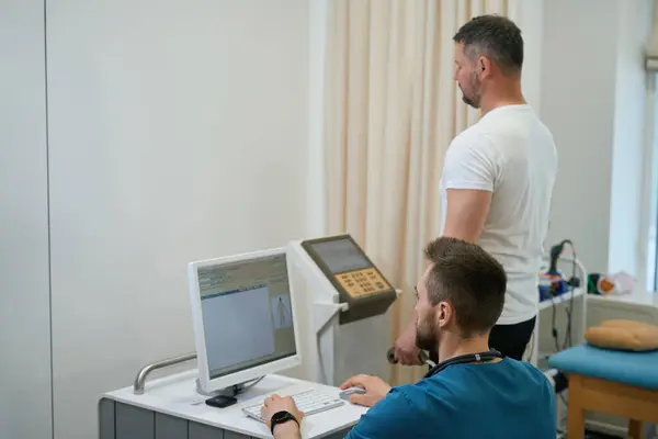 医生坐在办公桌前凝视着电脑显示屏 而病人则在分析仪上进行身体组成分析 — 图库照片