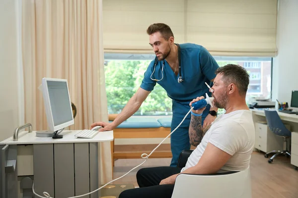 Patient with nose clip breathing into spirometer mouthpiece while pulmonologist monitoring his lung function on computer screen