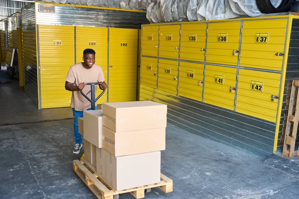 African American guy is pushing cargo cart around a warehouse, with cardboard boxes on the cart