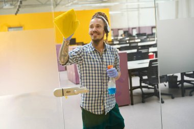 Koruyucu eldivenli neşeli bir adam camı sarı bir bezle ovuyor, temizleyici sprey kullanıyor.