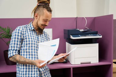 Rastalı bir adam fotokopi makinesinin yanında durur ve çalışma kâğıtlarını okur.