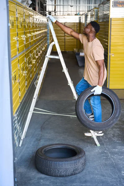 Guy Climbs Stepladder Car Tire His Hand Containers Yellow Warehouse — Stock Photo, Image