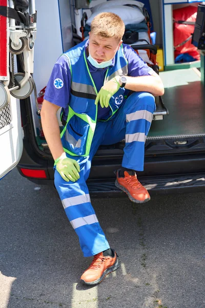 Young paramedic rests on the running board of a car between IVs, injections and patient procedures
