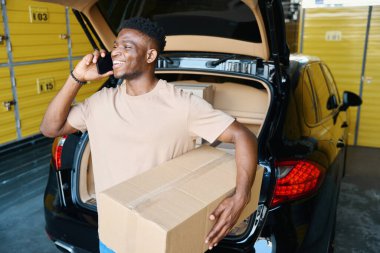 Afrikalı Amerikalı adam araba bagajını boşaltıp telefonla konuşuyor. Depoya bir şeyler getirmiş.