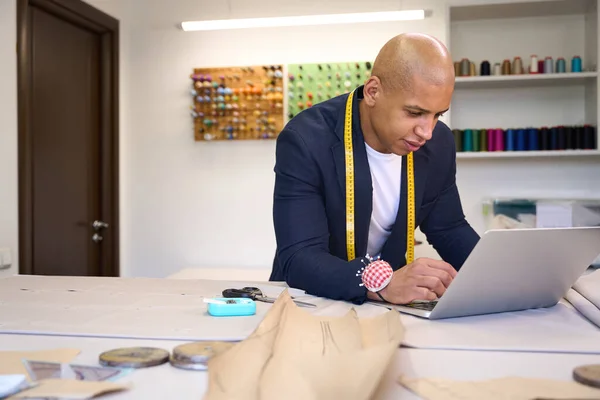 Fashion designer looking at laptop screen while leaning on cutting table in his atelier