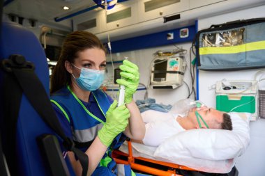 Ambulanstaki saha çalışanı şırıngayı ilaçla hazırlar, hasta oksijen maskesi ile yatar.