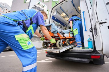 Paramedic in a protective mask unloads a patient from an ambulance, his colleague is near the patient clipart