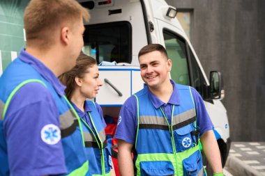Sağlık görevlileri ve hemşire otoparkta iletişim kuruyor. Modern bir binanın önünde bir ambulans bekliyor.