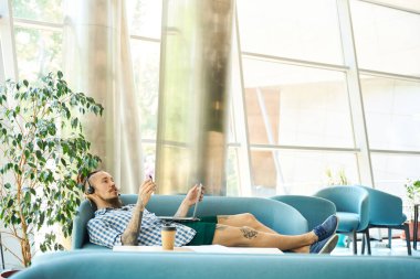 Genç serbest çalışan, çalışma alanındaki bir kanepede yatıyor, modern bir dizüstü bilgisayar ve kulaklık kullanıyor.