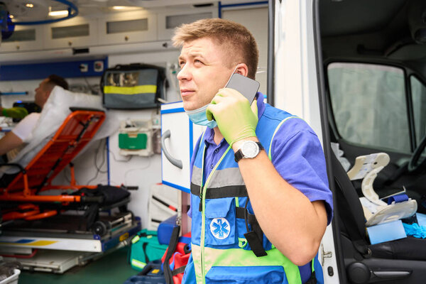 Paramedic communicates on a mobile phone near an ambulance, a patient lies on a gurney in the car
