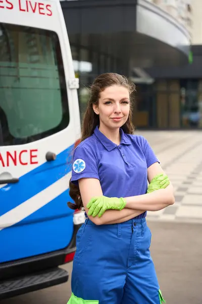 Female paramedic stands on the street near an ambulance, she has a medical emblem on her sleeve