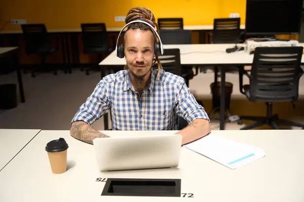 平らなシャツの非公式な男は オフィスデスクでラップトップと座って 男は仕事のためにヘッドセットを使用しています — ストック写真