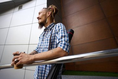 Modern bir binanın önünde elinde bir bardak kahveyle duran mutlu adamın çizimler için bir tüpü var.