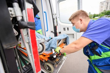 Bir hastayla birlikte özel bir sedyeyi modern bir ambulansa yükleyen sağlık görevlileri koruyucu eldivenlerle çalışıyorlar.