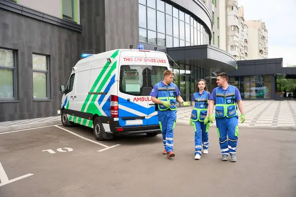 People in medical uniforms walk along the parking lot near a modern building, with an ambulance nearby