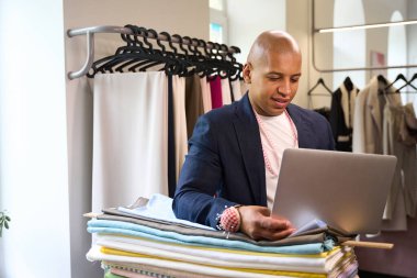 Gülümseyen usta terzi dirseğini kumaş rulolarına yaslarken dizüstü bilgisayara bakıyor.