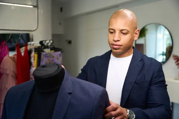 Concentrated African American tailor putting suit jacket on mannequin in his atelier