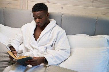 Afro-Amerikalı bir adam, rahat bir yatak odasında kitap okuyor, büyük bir yatakta oturuyor.