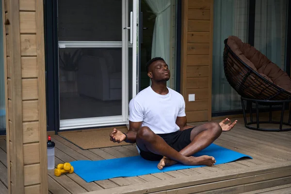 African American man practices yoga on a wooden terrace, he sits on a karimat