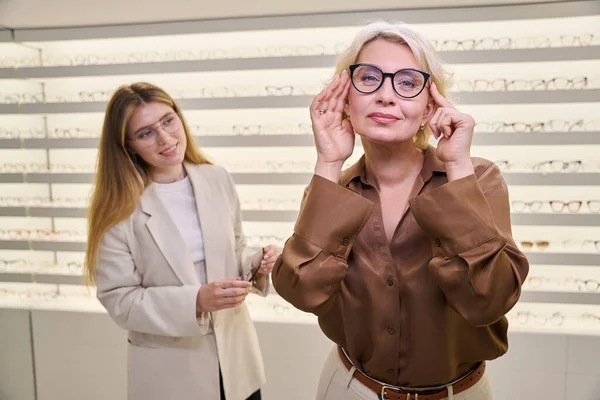 Charming middle-aged lady tries on glasses in an optical salon, with an ophthalmologist consultant nearby