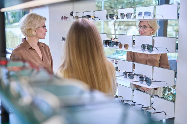 Middle-aged blonde tries on glasses in an opticians salon, being consulted by a young woman