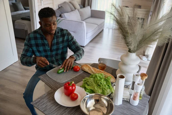Cute African American guy is preparing a salad of juicy vegetables, he is in comfortable casual clothes
