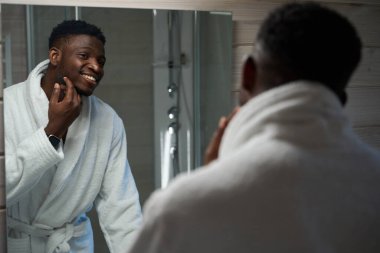 Erkek, banyoda aynada kendine bakar, rahat bir bornoz giyer.