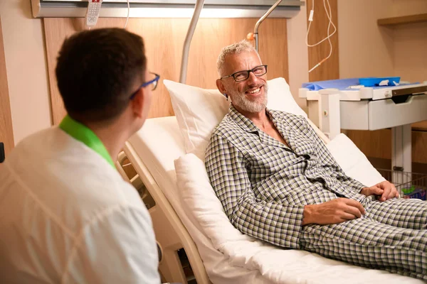 Smiling old man communicates with a doctor in the ward, near the bed there is an injection table