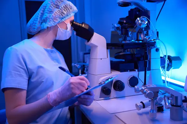 Female embryologist seated at laboratory table examining blastocysts through microscope while taking notes on clipboard