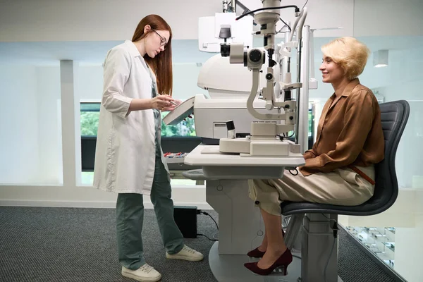 Red-haired doctor is consulting a middle-aged lady at diagnostic center, there are modern ophthalmological devices in the room