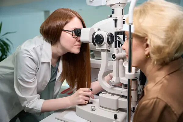女性眼科医は彼女の仕事で現代装置を使用し 顧客は機械の反対に座ります — ストック写真