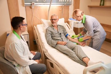 Bakıcı hemşire, yanında doktor olan gri saçlı yaşlı bir adama serum takıyor.