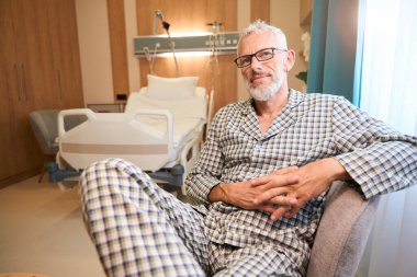 Tatmin olmuş hasta rahat bir odada ameliyattan sonra dinleniyor. Yumuşak bir sandalyede oturuyor.