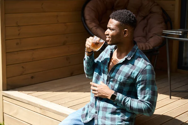 African American male on a wooden terrace with a cup of tea, he enjoys the summer morning