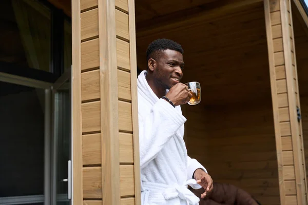 African American man greets morning on wooden terrace with a cup of tea, he is in a cozy terry robe