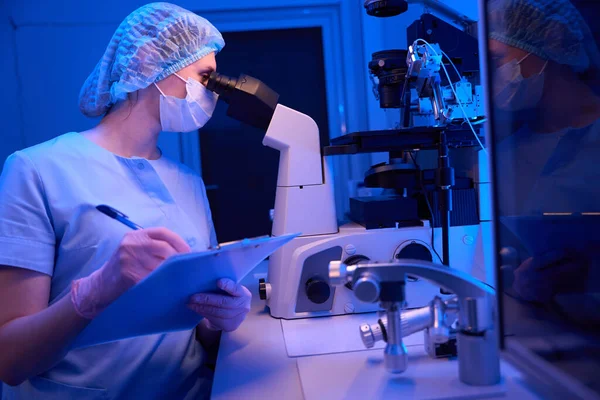 Female embryologist seated at laboratory table looking at specimens through microscope while writing on clipboard