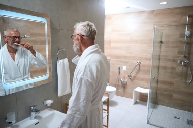 Neşeli yaşlı adam banyoda dişlerini fırçalıyor, tüylü bir bornoz giyiyor.