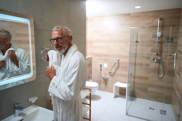 Old man in bathrobe wipes his face with towel in front of a mirror, he has a beard and glasses