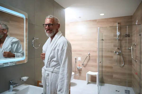 Old man in a bathrobe in the bathroom in front of a mirror, he has a beard and glasses