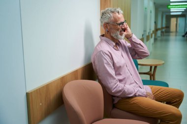 Gri saçlı adam hastane koridorunda cep telefonuyla iletişim kuruyor. Oda aydınlık ve temiz.