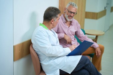 Erkekler parlak bir hastane koridorunda iletişim kuruyor, doktorun mavi bir tableti var.