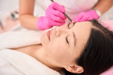 Bir kadın kaşlarını ameliyattan önce işaretleyen bir uzman kahverengi kalem kullanır.