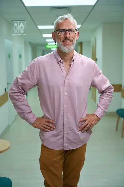 Man in glasses in a pink shirt stands in a hospital corridor, posing at camera