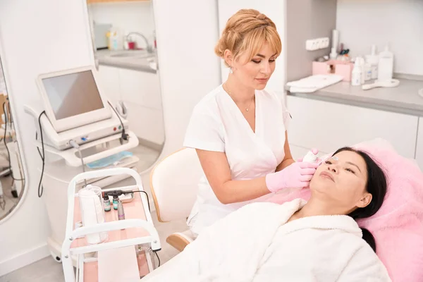 Beauty master applies cream to woman eyebrows, modern equipment indoors