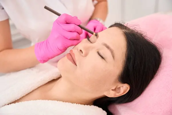Marking a woman eyebrows before the procedure, the master uses a brown pencil